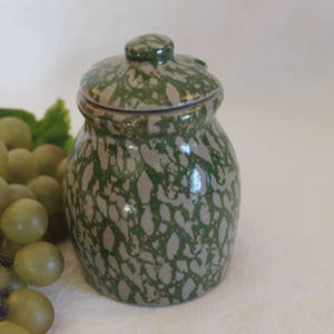 May include: A small ceramic jar with a lid, featuring a speckled green and grey glaze. The jar has a rounded shape and a slightly flared lip.