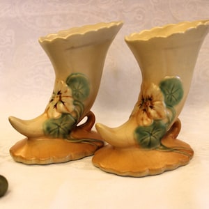 Peut inclure: Deux vases en céramique en forme de corne d'abondance vintage avec un motif floral. Les vases sont de couleur beige clair avec des feuilles vertes et des fleurs blanches.