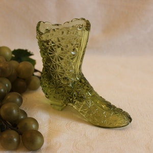 May include: A green glass boot-shaped vase with a diamond pattern. The vase is sitting on a white surface.