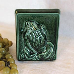 May include: A green ceramic tile with a raised image of two hands clasped in prayer.
