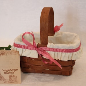 May include: A small, brown wicker basket with a white fabric lining and pink ribbon accents. The basket has a wooden handle and is sitting on a white surface. The Longaberger Baskets logo is visible on a tag in the foreground.