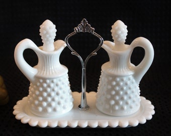 May include: White milk glass oil and vinegar cruets with a silver metal stand. The cruets have a textured, dotted surface and a small spout.