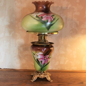 May include: A green and brown glass oil lamp with a floral design. The lamp has a round shade with a scalloped edge and a base with three feet.