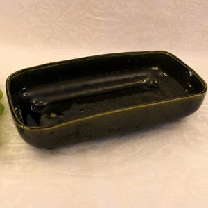 May include: A dark green ceramic rectangular dish with a speckled glaze. The dish has a slightly raised rim and three small feet.