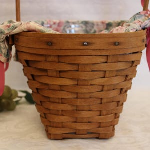 May include: A brown wicker basket with a pink and floral fabric lining. The basket has a handle and is decorated with metal rivets.