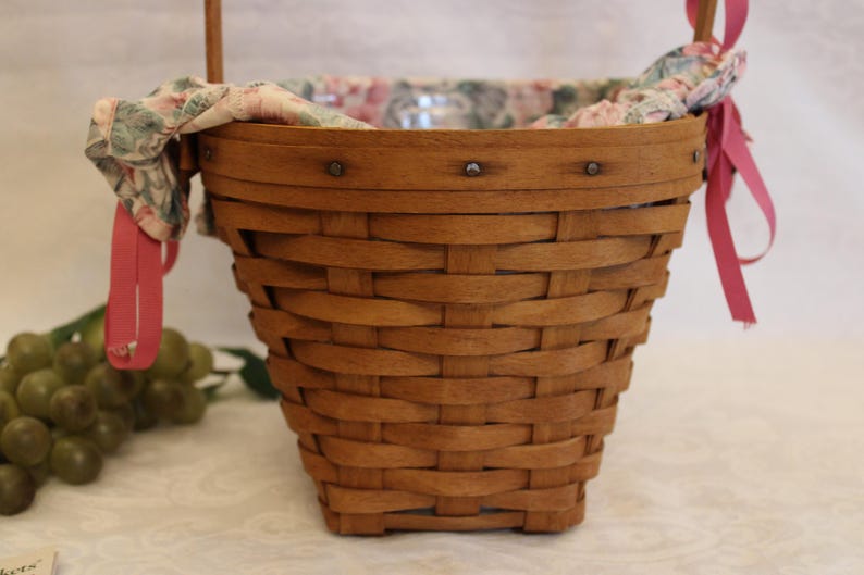 May include: A brown wicker basket with a pink and floral fabric lining. The basket has a handle and is decorated with metal rivets.