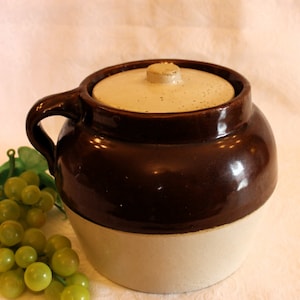 May include: A brown and white glazed ceramic crock with a lid. The crock has a handle and is sitting on a white surface.