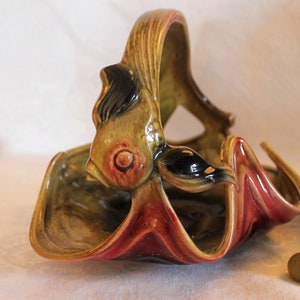 May include: A ceramic fruit bowl shaped like a fish with a green, black, and red glaze. The bowl has a handle that is part of the fish's body.