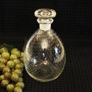 May include: A clear glass bottle with a textured surface and a round stopper. The bottle is shaped like a teardrop and has a small opening at the top.