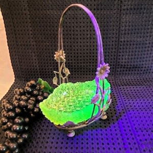 May include: A vintage green glass basket with a metal handle and floral accents. The basket has a decorative pattern and sits on a metal base. A bunch of dark grapes is next to the basket.