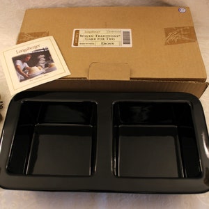 May include: Black ceramic baking dish with two divided sections. The dish is in its original box, which has a label that reads "Longaberger Woven Traditions Cake for Two Ebony".