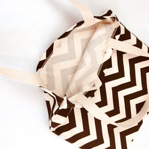 K&ouml;nnte beinhalten: Eine braun-cremefarbene Tragetasche mit Chevron-Muster und cremefarbenen Griffen. Die Tasche ist offen und zeigt das Innenfutter. Die Tasche ist aus Stoff und eignet sich zum Tragen von Gegenst&auml;nden.