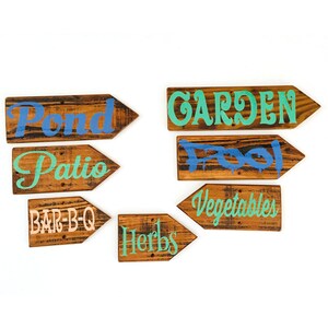 May include: Wooden arrow signs with various destinations painted in different colors. Destinations include "Pond", "Patio", "Bar-B-Q", "Garden", "Pool", "Vegetables", and "Herbs". The signs are made of wood with a distressed finish.