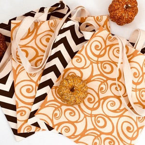 K&ouml;nnte beinhalten: Vier Canvas-Tragetaschen mit cremefarbenen Griffen. Zwei Taschen haben ein braunes und cremefarbenes Zickzackmuster, und zwei haben einen cremefarbenen Hintergrund mit einem braunen Wirbeldesign. Dekorative K&uuml;rbisse und ein Bagel sind in der N&auml;he der Taschen platziert.