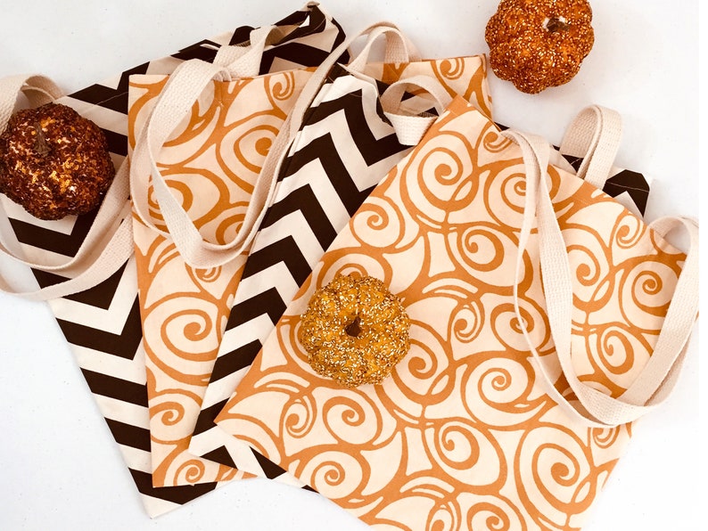 K&ouml;nnte beinhalten: Vier Canvas-Tragetaschen mit cremefarbenen Griffen. Zwei Taschen haben ein braunes und cremefarbenes Zickzackmuster, und zwei haben einen cremefarbenen Hintergrund mit einem braunen Wirbeldesign. Dekorative K&uuml;rbisse und ein Bagel sind in der N&auml;he der Taschen platziert.