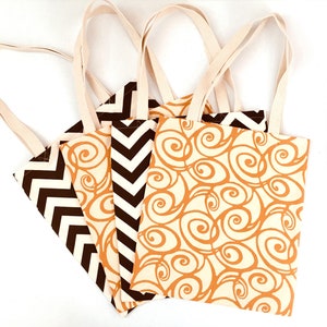 K&ouml;nnte beinhalten: Vier Tragetaschen aus Canvas mit cremefarbenen Griffen. Die Taschen weisen verschiedene Muster auf: ein braunes und cremefarbenes Chevron, ein braunes und cremefarbenes Wirbeldesign und ein braunes und cremefarbenes Zickzackmuster. Die Taschen sind auf einer wei&szlig;en Oberfl&auml;che angeordnet.