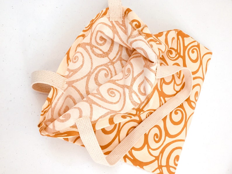 K&ouml;nnte beinhalten: Beige Tragetasche mit einem sich wiederholenden Muster aus wirbelnden, rostfarbenen Designs. Die Tasche hat cremefarbene Griffe und ist aus einem weichen Stoff gefertigt. Auch das Innere der Tasche ist sichtbar und zeigt das gleiche Muster.
