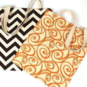 K&ouml;nnte beinhalten: Zwei Tragetaschen aus Canvas mit cremefarbenen Henkeln. Eine Tasche hat ein braunes und cremefarbenes Zickzackmuster, die andere ein oranges Wirbeldesign auf cremefarbenem Hintergrund. Beide Taschen sind auf wei&szlig;em Hintergrund.