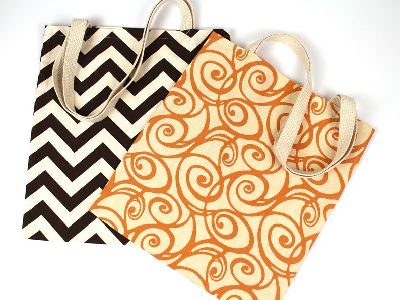 K&ouml;nnte beinhalten: Zwei Tragetaschen aus Canvas mit cremefarbenen Henkeln. Eine Tasche hat ein braunes und cremefarbenes Zickzackmuster, die andere ein oranges Wirbeldesign auf cremefarbenem Hintergrund. Beide Taschen sind auf wei&szlig;em Hintergrund.