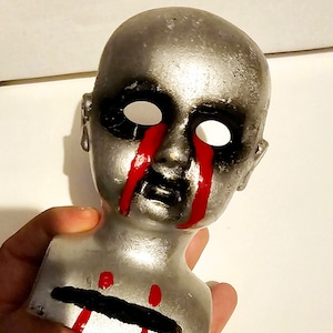 May include: A silver doll's head with black eyes and red paint dripping down the face. The doll's head is on a silver base.
