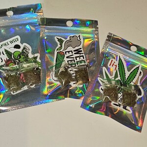May include: Three small, clear plastic bags containing dried green plant material. Each bag is sealed and placed inside a holographic, iridescent pouch. Stickers with cannabis-related graphics and text, including "SMOKE WEED" and "WEED EVER", are affixed to the pouches.