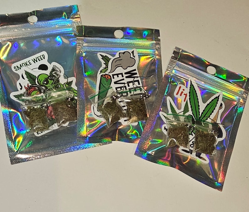 May include: Three small, clear plastic bags containing dried green plant material. Each bag is sealed and placed inside a holographic, iridescent pouch. Stickers with cannabis-related graphics and text, including "SMOKE WEED" and "WEED EVER", are affixed to the pouches.