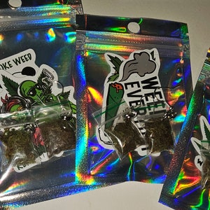 May include: Several small, clear plastic bags filled with dried herbs are visible inside iridescent, holographic pouches. Each pouch has a sticker with a cannabis-related design and text. The bags are sealed with a small metal clasp.