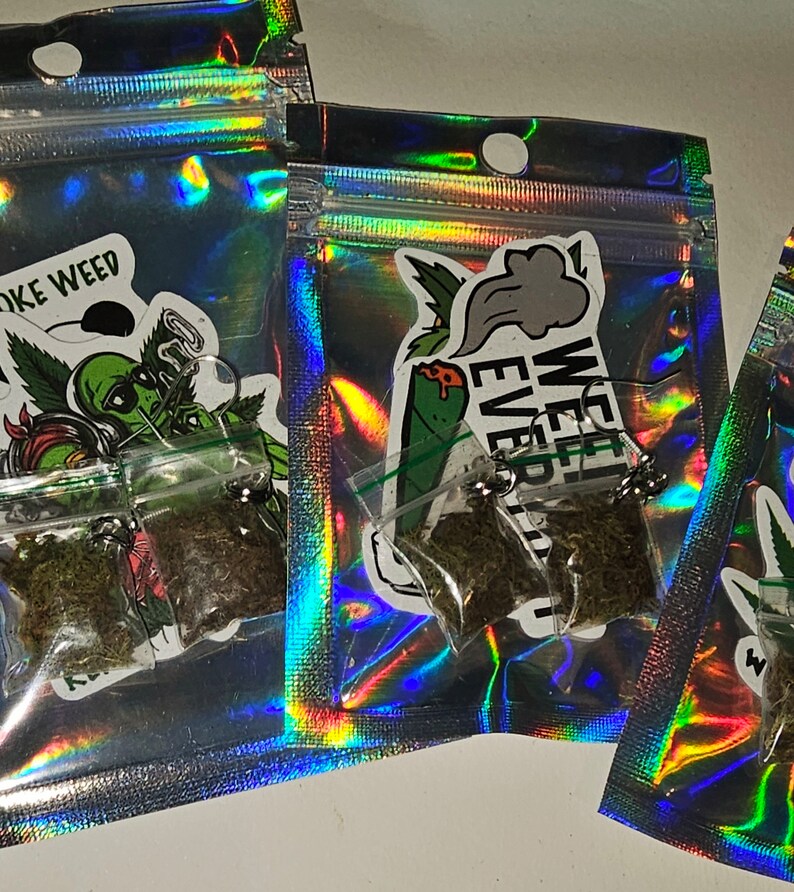 May include: Several small, clear plastic bags filled with dried herbs are visible inside iridescent, holographic pouches. Each pouch has a sticker with a cannabis-related design and text. The bags are sealed with a small metal clasp.