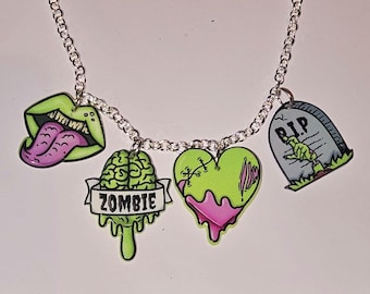 Pode incluir: Um colar de corrente prateada com cinco pingentes: uma língua verde saindo de uma boca, um cérebro verde com a palavra "ZOMBIE" em uma faixa, um coração verde com bordas rosa pingando e uma lápide cinza com as palavras "RIP" e uma mão verde.
