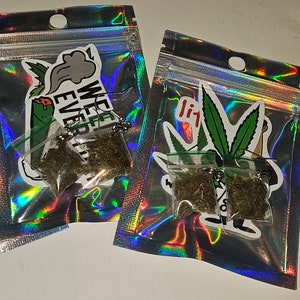 May include: Two small clear plastic bags filled with dried green herbs, attached to silver keychains. The bags are inside iridescent, holographic pouches. Stickers with cannabis leaf designs and text are visible on the pouches. One sticker says "WE EVER".