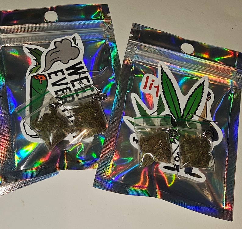 May include: Two small clear plastic bags filled with dried green herbs, attached to silver keychains. The bags are inside iridescent, holographic pouches. Stickers with cannabis leaf designs and text are visible on the pouches. One sticker says "WE EVER".