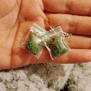 May include: A pair of novelty earrings featuring small plastic bags filled with green and brown plant material. The earrings have silver-colored ear wires and are held in a hand against a fuzzy, light-colored background.