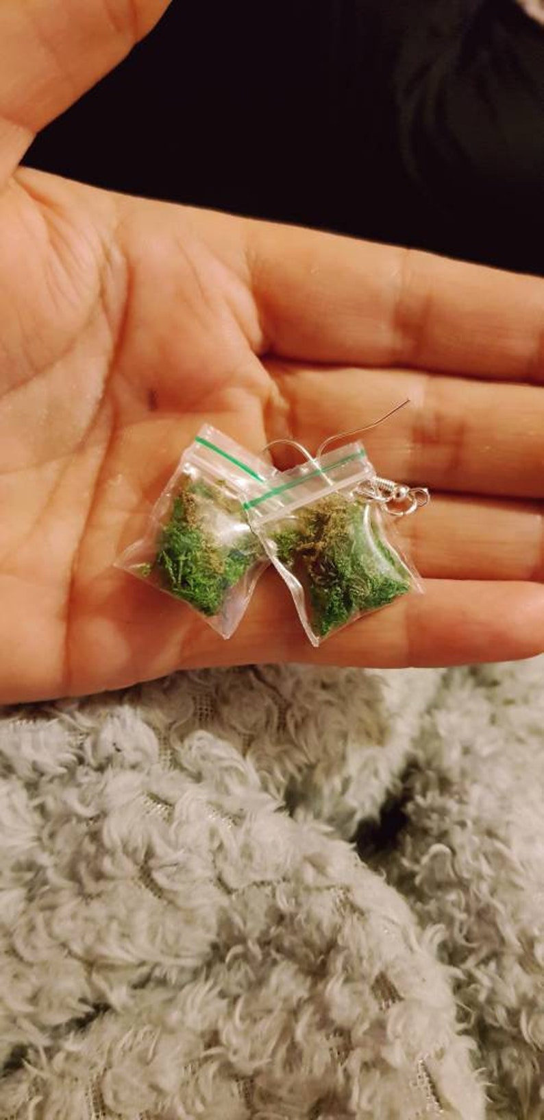 May include: A pair of novelty earrings featuring small plastic bags filled with green and brown plant material. The earrings have silver-colored ear wires and are held in a hand against a fuzzy, light-colored background.