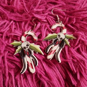 May include: A pair of silver dangle earrings with a white candy cane design. The candy cane is decorated with red and white stripes and has a green holly leaf and a skull with a black bow tie.
