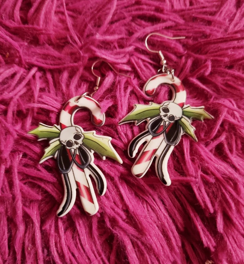May include: A pair of silver dangle earrings with a white candy cane design. The candy cane is decorated with red and white stripes and has a green holly leaf and a skull with a black bow tie.