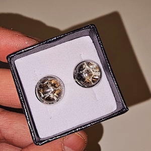 Op de afbeelding: Een paar zilveren manchetknopen met een helder glazen centrum met een bruin en gouden ontwerp. De manchetknopen worden gepresenteerd in een zwarte fluwelen doos.