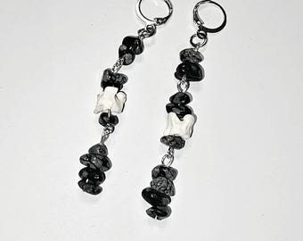 Pode incluir: Um par de brincos pendurados com contas de pedra salpicadas em preto e branco e pingentes brancos em forma de osso. Os brincos têm fechos de alavanca prateados e são exibidos em um fundo branco.