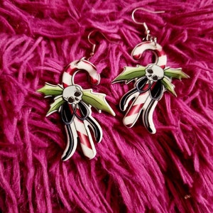 May include: A pair of silver dangle earrings with a candy cane and skull design. The candy cane is red and white with green holly leaves and a black skull.