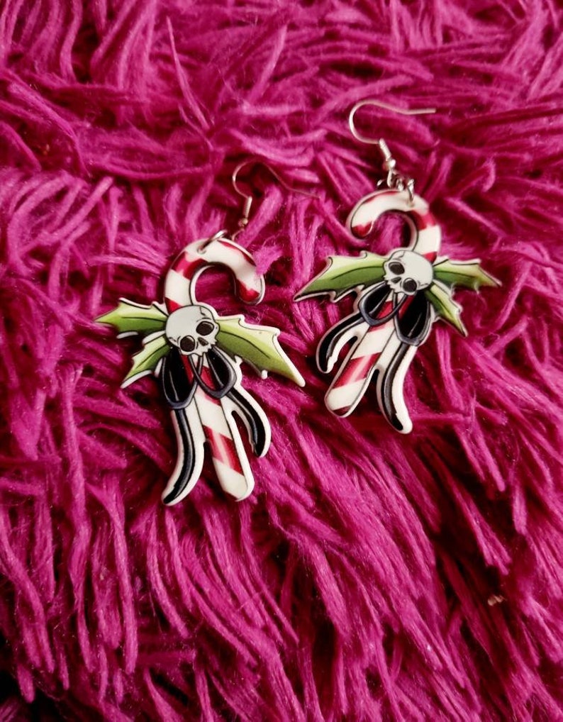 May include: A pair of silver dangle earrings with a candy cane and skull design. The candy cane is red and white with green holly leaves and a black skull.