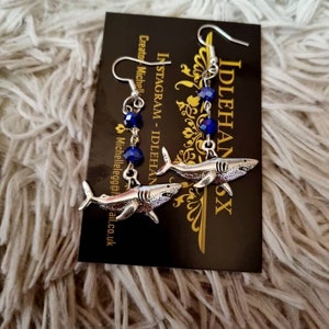 Pode incluir: Um par de brincos de tubar&atilde;o prateados com contas de cristal azul. Os brincos est&atilde;o pendurados em um cart&atilde;o que diz "IdlehandsX" e "Instagram - IdlehandsX".