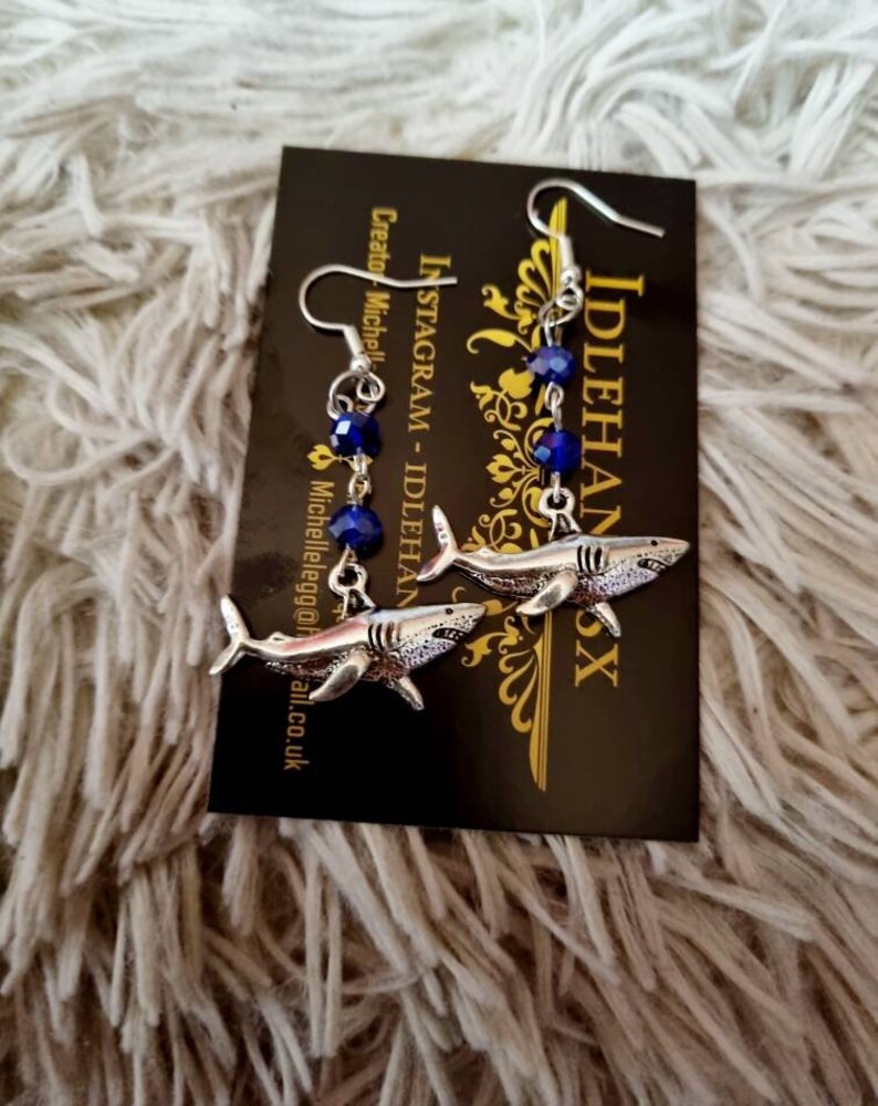 Pode incluir: Um par de brincos de tubar&atilde;o prateados com contas de cristal azul. Os brincos est&atilde;o pendurados em um cart&atilde;o que diz "IdlehandsX" e "Instagram - IdlehandsX".