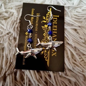 Pode incluir: Um par de brincos de tubar&atilde;o prateados com contas de cristal azul penduradas nos pingentes de tubar&atilde;o. Os brincos est&atilde;o em um cart&atilde;o preto com o texto "IdleHandsX" e "Instagram - IdleHandsX Creator - Michelle" impresso nele.