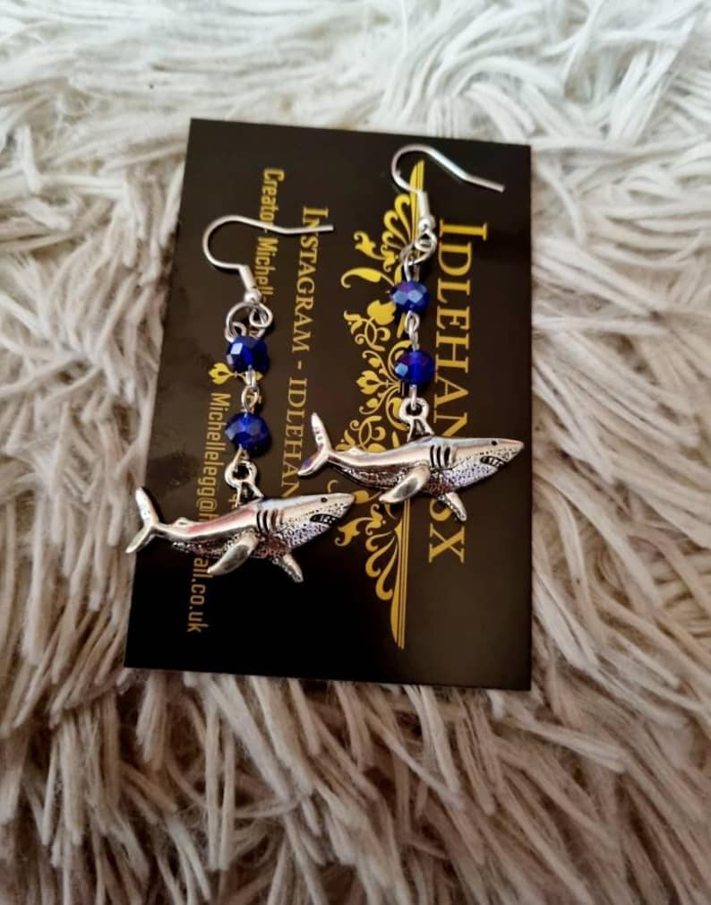 Pode incluir: Um par de brincos de tubar&atilde;o prateados com contas de cristal azul penduradas nos pingentes de tubar&atilde;o. Os brincos est&atilde;o em um cart&atilde;o preto com o texto "IdleHandsX" e "Instagram - IdleHandsX Creator - Michelle" impresso nele.