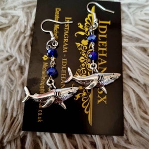 Pode incluir: Brincos de tubar&atilde;o prateados com contas de cristal azul. Os brincos est&atilde;o pendurados em um cart&atilde;o preto com o texto "IDLEHANDSX" e "INSTAGRAM - IDLEHANDSX Creator: Michelle"