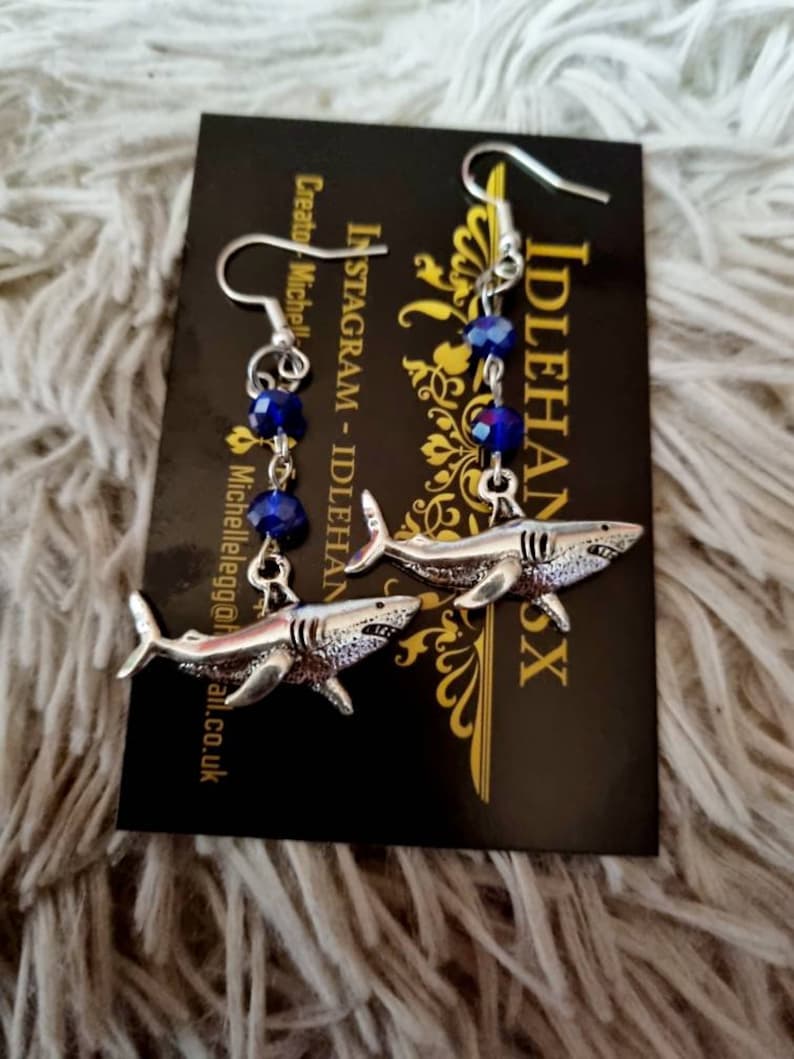 Pode incluir: Brincos de tubar&atilde;o prateados com contas de cristal azul. Os brincos est&atilde;o pendurados em um cart&atilde;o preto com o texto "IDLEHANDSX" e "INSTAGRAM - IDLEHANDSX Creator: Michelle"