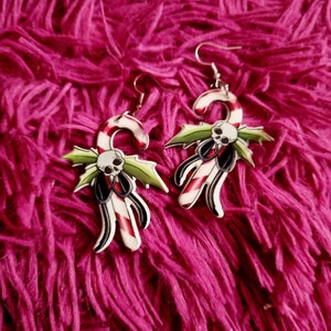 May include: A pair of silver earrings shaped like candy canes with a skull and holly design. The candy canes are red and white with black accents.