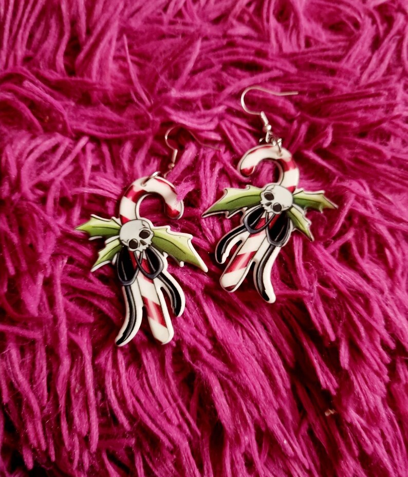 May include: A pair of silver earrings shaped like candy canes with a skull and holly design. The candy canes are red and white with black accents.