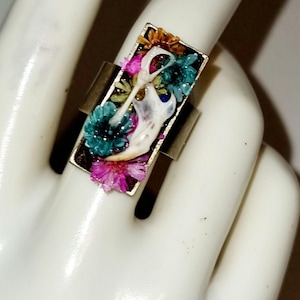 Puede incluir: Un anillo de plata con una incrustación rectangular de resina que presenta un hueso blanco y flores prensadas de colores. Las flores son de tonos rosa, azul verdoso, amarillo y naranja.