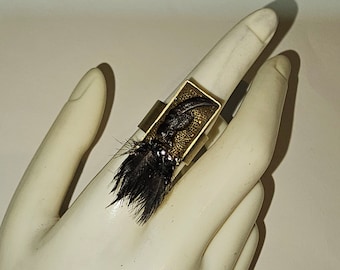May include: A gold ring with a black feather and a rectangular, gold-toned, textured piece of metal with a dark brown bird claw inside.