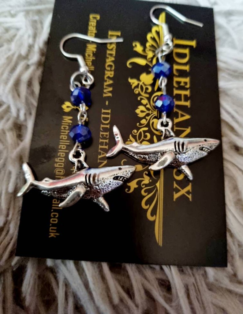 Pode incluir: Brincos de tubar&atilde;o prateados com contas de cristal azul. Os brincos est&atilde;o pendurados em um cart&atilde;o preto com o texto "INSTAGRAM - IDLEHANDSX" nele.